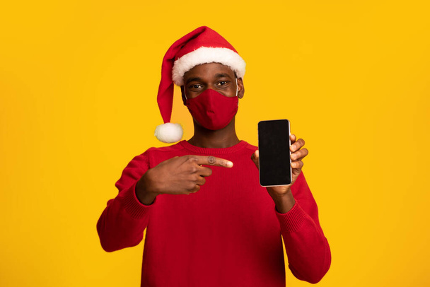 Africano chico en máscara y sombrero de Santa señalando en la pantalla del teléfono inteligente negro - Foto, Imagen