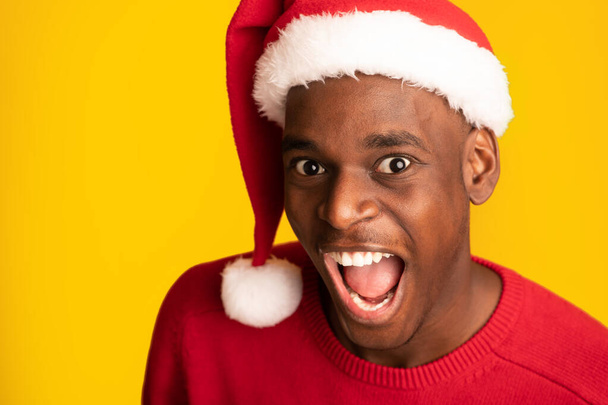 Detailní záběr šílené emocionální černošky chlap v Santa klobouku přes žluté pozadí - Fotografie, Obrázek
