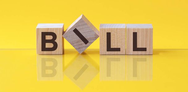 Билл слово написано на деревянном блоке. Содержание слова состоит из деревянных строительных блоков, лежащих на желтом столе. Законопроект, бизнес-концепция, желтый фон - Фото, изображение