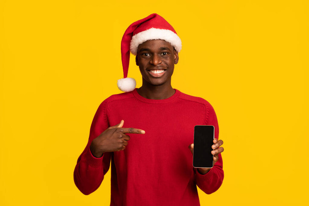Θετικός Αφρικανός με καπέλο Σάντα δείχνει στο Smartphone με μαύρη οθόνη - Φωτογραφία, εικόνα