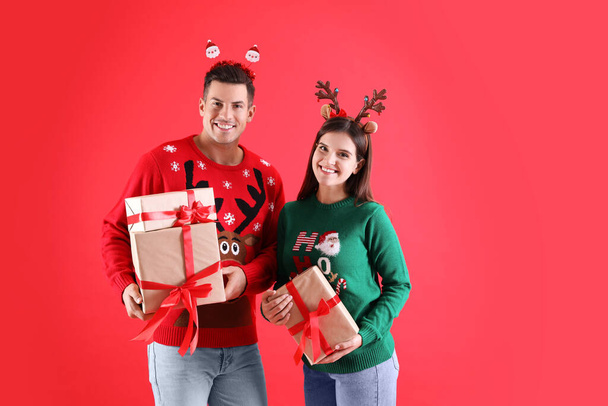 Beautiful happy couple in Christmas headbands and sweaters holding gifts on red background - Fotografie, Obrázek
