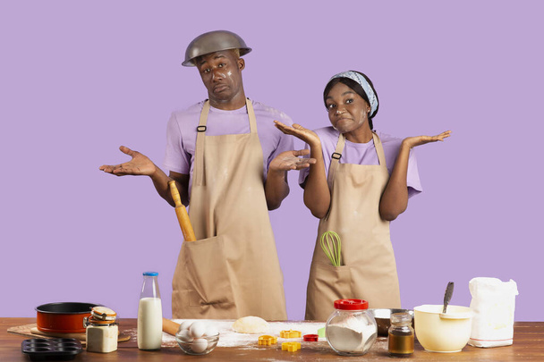 Смущенная черная пара, вся в муке, пожимающая плечами, не умеющая готовить, стоящая озадаченная на фоне фиолетовой студии - Фото, изображение