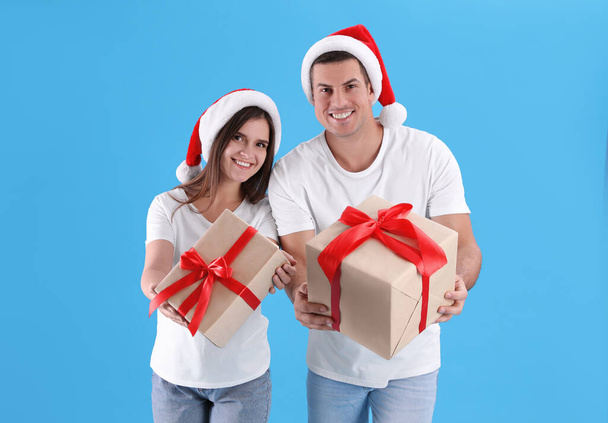 Beautiful happy couple in Santa hats holding Christmas gifts on light blue background - Foto, Bild