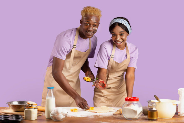 Joyful millennial couple enjoying homemade cooking, holding cookie cutters, baking together on lilac studio background - Photo, image