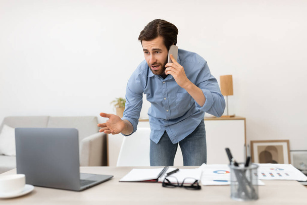 Unzufriedener Geschäftsmann spricht mit Handy und hat Computerproblem im Büro - Foto, Bild
