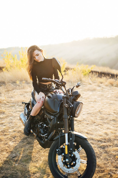 Beautiful girl in black clothes sits on motorcycle and poses for camera. Evening sunset - Zdjęcie, obraz