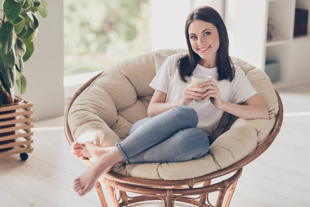 Full size photo of pretty girl sit on wicker chair hold hot tasty coffee cup in house living room - 写真・画像