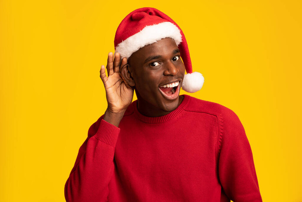zwarte man in kerstman hoed vasthouden hand in de buurt oor proberen afluisteren iets - Foto, afbeelding