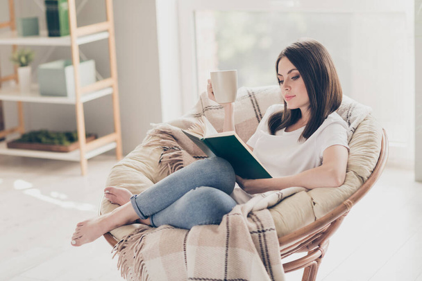Foto de comprimento total da menina concentrada sentar na cadeira de vime ler livro segurar xícara de café no quarto da casa moderna - Foto, Imagem