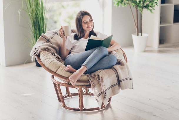 Full body photo of positive young girl sit wicker chair read book hold coffee cup wear white t-shirt bare foot in house indoors - Valokuva, kuva