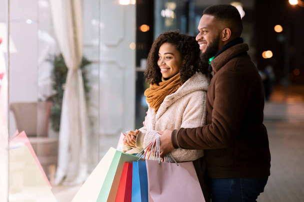 Feliz pareja negra con bolsas de compras mirando a la ventana del centro comercial - Foto, Imagen