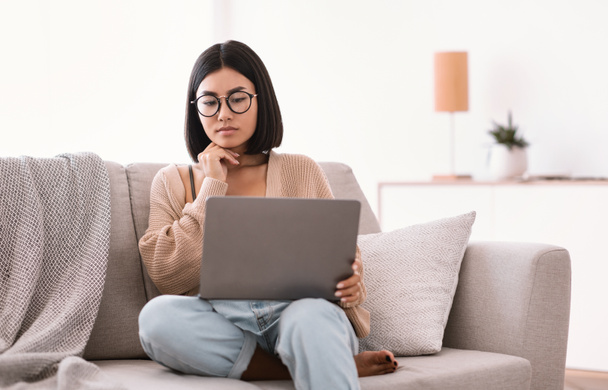 Asian woman sitting on couch, using laptop and thinking - Photo, Image