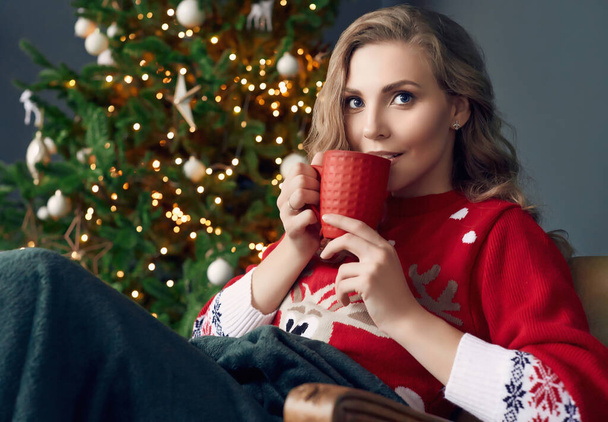 Ritratto di splendida donna bionda in maglione rosso che si rilassa sotto un plaid con una tazza di caffè nell'interno decorato di Natale. - Foto, immagini