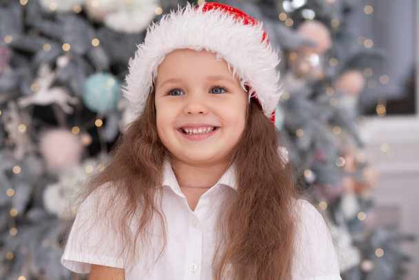 New Year's portrait of a girl, a cute child in a Santa Claus hat against the background of a New Year tree, Christmas mood, winter holiday, baby laughs. - Photo, Image