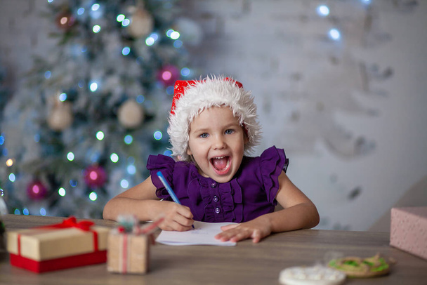 A cute little girl writes a New Year's letter to Santa Claus, the child is sitting at the table, a Christmas tree, a festive mood, childhood and happiness. - Foto, Imagen