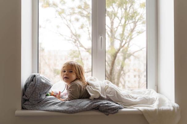 Child lying on the window in living room and watching at the street. cozy home on weekends, holidays and lockdown - Фото, изображение