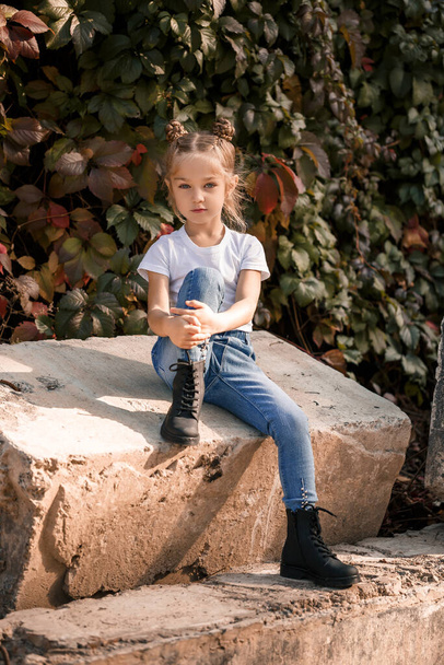 Street photo of a little beautiful girl in jeans and a white t-shirt on a background of concrete slabs and autumn leaves - Foto, Imagem