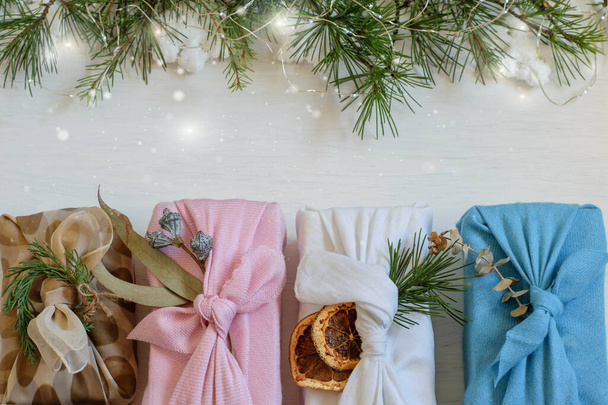Vánoční ručně vyráběné dárkové krabice zabalené v opětovném použití látky v japonském stylu furoshiki. Slavnostní bílé pozadí s vánoční strom větve, bokeh světla, kopírovací prostor. Balíček péče, diy pojmy. - Fotografie, Obrázek