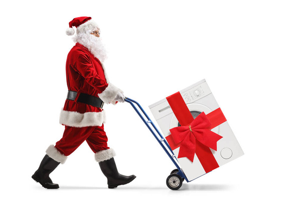 Full length portrait of a santa claus pushing a washing machine with a red bow on a hand truck isolated on white background - Fotó, kép