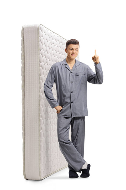 Full length portrait of a young male in pajamas leaning on a matress and pointing up isolated on white background - Foto, immagini