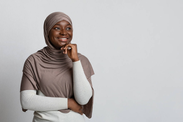 Заинтересованная чёрная женщина в хиджабе смотрит на копировальное пространство и трогает подбородок - Фото, изображение