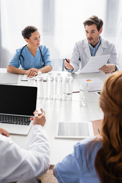 Doctor holding paper and talking to multiethnic colleagues near devices and water on table  - Photo, Image