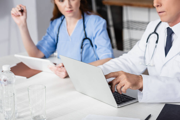 Cropped view of doctor pointing at laptop near nurse with digital tablet on blurred background  - Photo, Image
