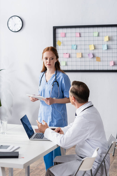 Enfermera con tableta digital mirando al médico cerca de la computadora portátil y portátil en primer plano borroso  - Foto, Imagen