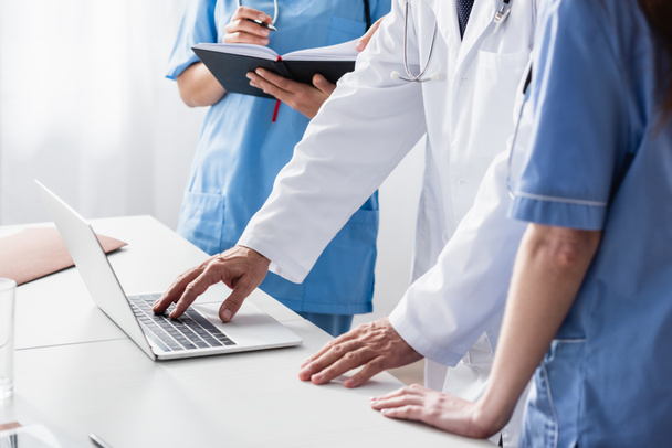 Vista recortada del médico en bata blanca usando portátil cerca de enfermeras con cuaderno  - Foto, imagen