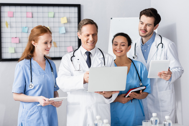 Fröhliche multiethnische Ärzte mit Geräten und Notizbuch im Krankenhaus  - Foto, Bild