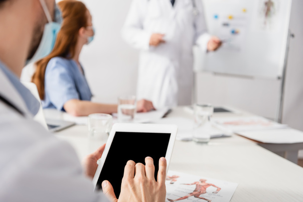 Doctor in medical mask using digital tablet with blank screen near document and colleagues on blurred background  - Photo, Image