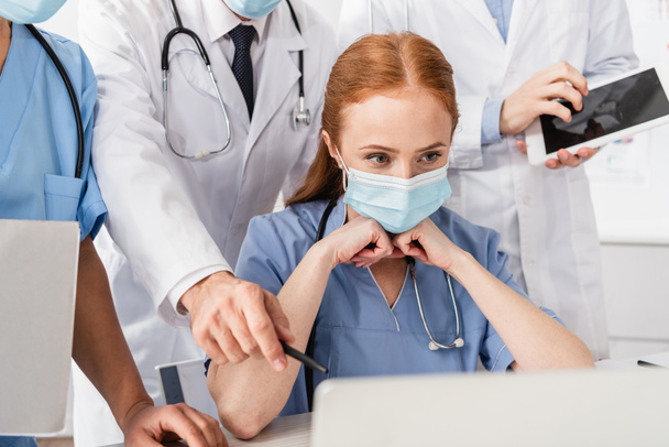 Руда медсестра в медичній масці дивиться на ноутбук біля мультикультурних колег, стоячи позаду на розмитому передньому плані
 - Фото, зображення