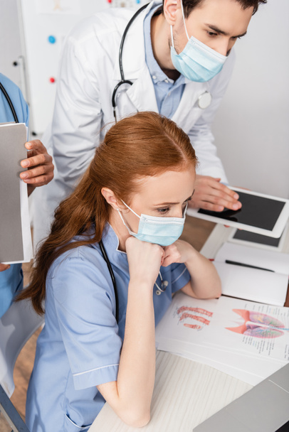 Infermiera in maschera medica seduta sul posto di lavoro vicino a colleghi multiculturali su sfondo sfocato - Foto, immagini