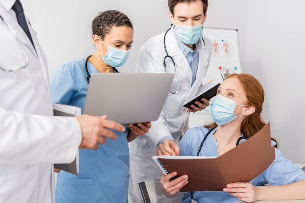 Enfermera en máscara médica sosteniendo la carpeta de papel mientras está sentada cerca de colegas multiétnicos en el hospital - Foto, Imagen