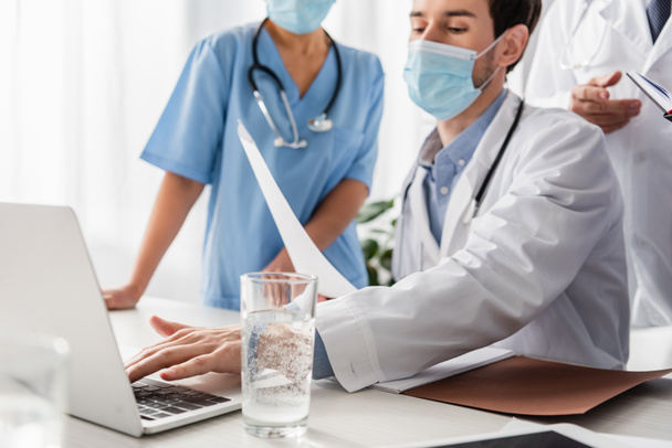 Doctor in medical mask with paper typing on laptop near multiethnic colleagues on blurred foreground in hospital - Photo, Image