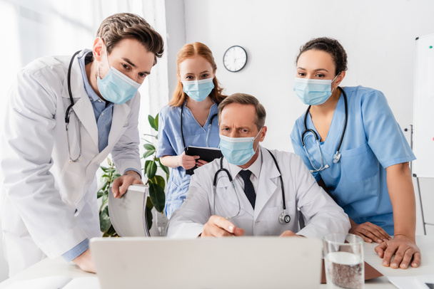 Médecins et infirmières multiethniques portant des masques médicaux utilisant un ordinateur portable sur le lieu de travail à l'hôpital au premier plan flou - Photo, image