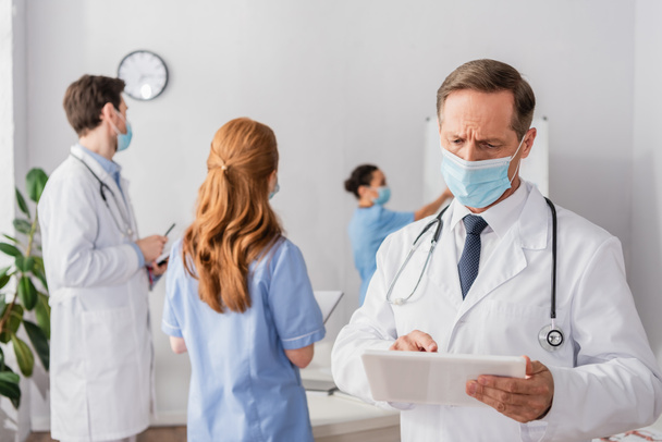 Doctor in medical mask using digital tablet with blurred multiethnic colleagues on background - Photo, Image