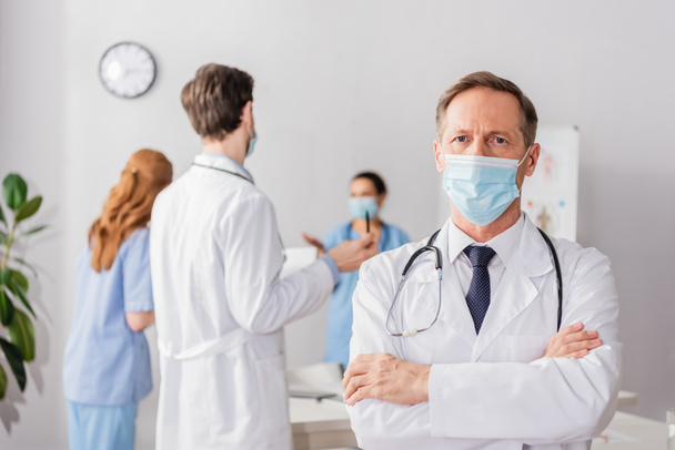 Médico em máscara médica com braços cruzados olhando para a câmera com colegas borrados trabalhando no fundo - Foto, Imagem
