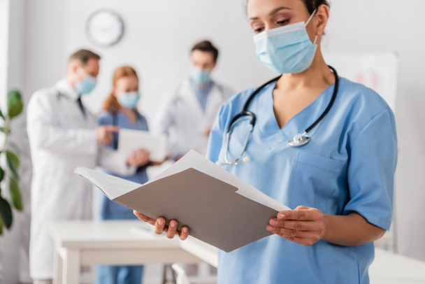 African american nurse looking at paper folder with blurred colleagues on background - Photo, Image