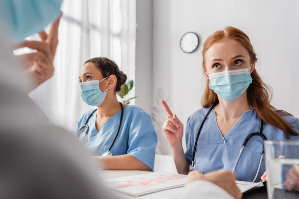 Redhead νοσοκόμα δείχνει προειδοποιητική χειρονομία, ενώ κάθεται με πολυπολιτισμικούς συναδέλφους στο χώρο εργασίας σε θολή πρώτο πλάνο - Φωτογραφία, εικόνα