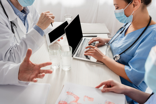 African american nurse typing on laptop near colleagues working at workplace with tablet, papers and glasses of water on blurred foreground - Photo, Image