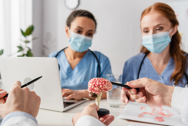 Arts wijzend met pen op hersenen anatomisch model op de werkplek met wazig multi-etnische verpleegkundigen op de achtergrond in het ziekenhuis - Foto, afbeelding