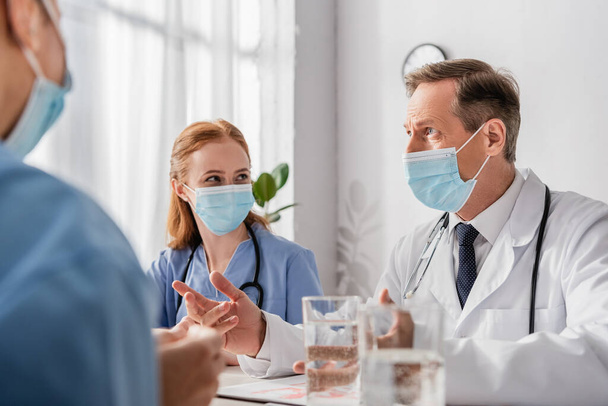 Doctor in medical mask talking to colleagues while sitting at workplace with blurred glasses of water on foreground - Fotografie, Obrázek