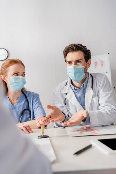 Orvos orvosi maszk beszél kollégáival, miközben ül a munkahelyen papírok és az agy anatómiai modell elmosódott előtérben - Fotó, kép