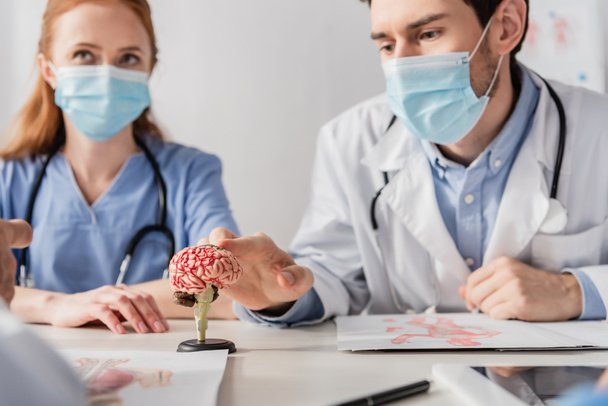 Doctor in medical mask touching brain anatomical model while sitting at workplace near colleagues on blurred foreground - Photo, Image