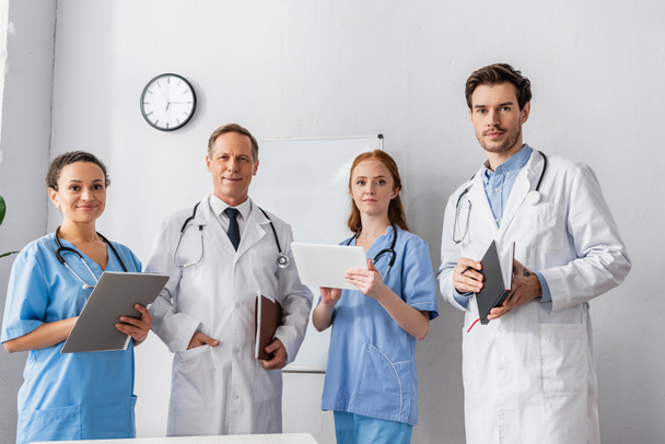 Pozitív többnemzetiségű kórházi személyzet papírokkal és digitális táblagéppel, flipchart és falióra a háttérben - Fotó, kép