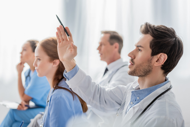 Arzt mit erhobener Hand neben multiethnischen Kollegen während eines Treffens im Krankenhaus auf verschwommenem Hintergrund - Foto, Bild