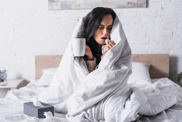 sick young brunette woman with hot drink sneezing in bed - Photo, Image