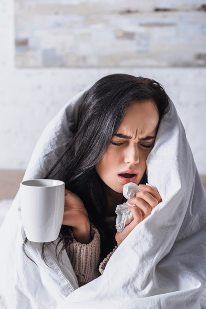 kranke junge brünette Frau mit Heißgetränk niest im Bett - Foto, Bild