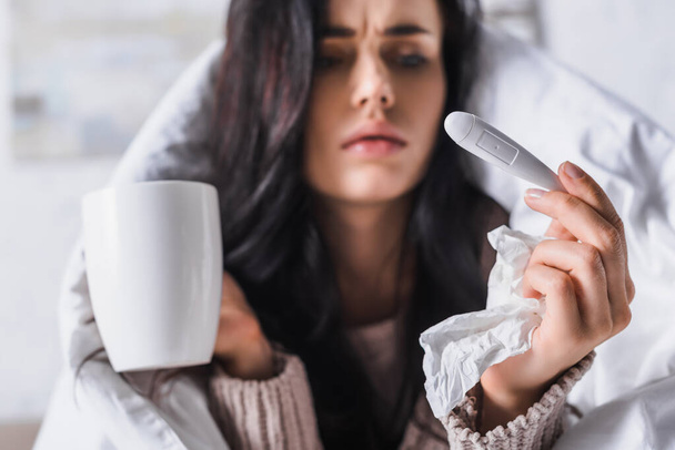 kranke junge brünette Frau mit Gewebe, Thermometer und Heißgetränk im Bett - Foto, Bild